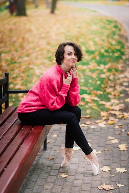 Młoda piękna balerina w jasnym płaszczu i pointe siedzieć na ławce, odpoczywać na świeżym powietrzu w jesiennym parku.