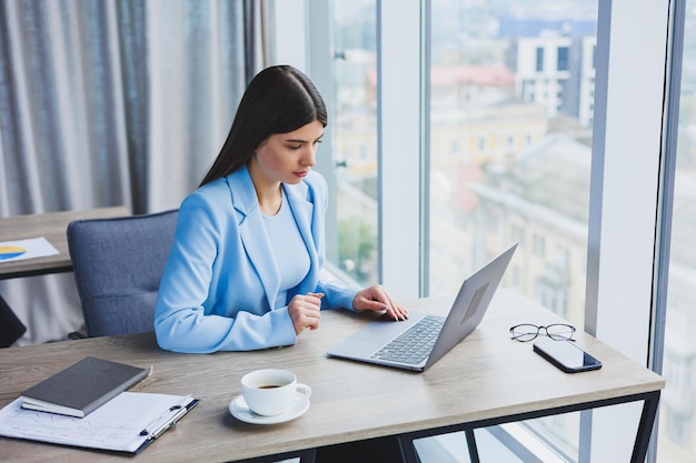 Młoda piękna biznesowa kobieta w niebieskiej koszuli siedzi w biurze z laptopem i robi interesy