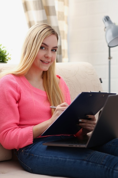 młoda piękna blond kobieta siedzieć na kanapie w salonie trzymać spoiwo w pracy na laptopie