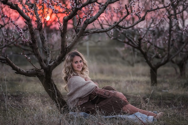 Młoda piękna blondynka kręcona w brązowej plisowanej spódnicy, różowej bluzce, zakryte ramiona z dzianinowym szalikiem, siedzi w kwitnących brzoskwiniowych ogrodach o zachodzie słońca