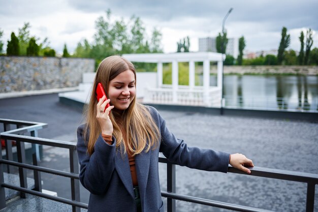 Młoda piękna blondynka w szarym płaszczu rozmawia przez telefon w zimny jesienny dzień. Dziewczyna z telefonem na tarasie z widokiem na rzekę