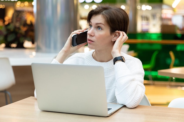 Młoda piękna brunetka kobieta siedzi w centrum handlowym przy stole i pracuje na laptopie przy użyciu telefonu komórkowego. Freelancer i koncepcja biznesowa