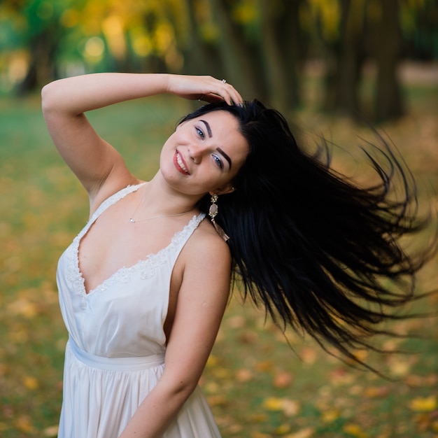 Młoda piękna brunetka kobieta w długiej białej sukni w parku jesienią.