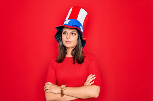 Młoda piękna brunetka kobieta w kapeluszu stanów zjednoczonych świętuje dzień niepodległości, patrząc z boku ze skrzyżowanymi rękami przekonana i pewna siebie