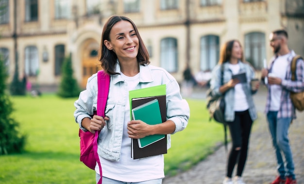 Młoda Piękna Brunetka Studentka W Dżinsowych Ubraniach Z Plecakiem I Książkami W Rękach Stoi Na Grupie Jej Przyjaciół Studentów