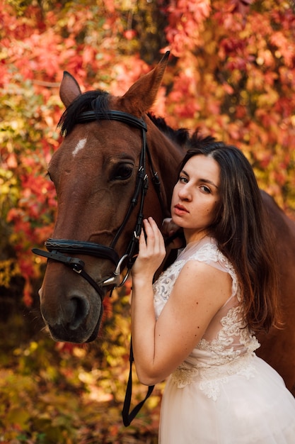 Młoda piękna brunetka w białej sukni przytula brązowego konia