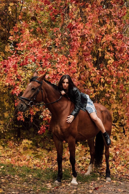 Młoda piękna brunetka w sukience i skórzanej czarnej kurtce seksownie leży na koniu