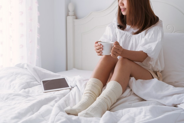 Młoda piękna brunetka włosy kobieta z filiżanką kawy, siedząc na łóżku przy oknie.