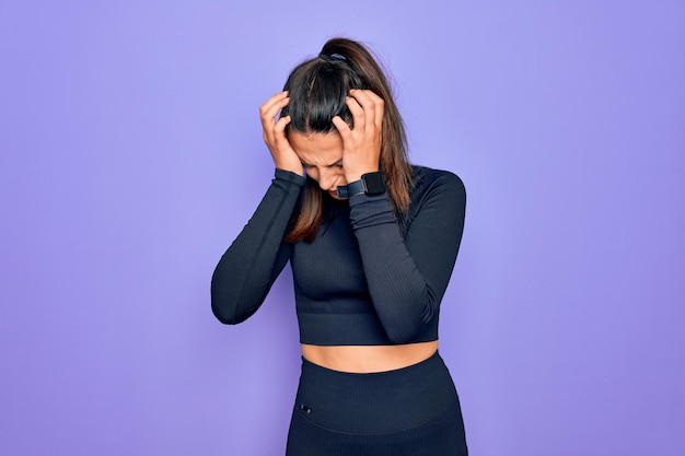 Młoda piękna brunetka wysportowana kobieta ubrana w swobodną odzież sportową na fioletowym tle cierpiąca na ból głowy zdesperowana i zestresowana z powodu bólu i migreny Ręce na głowie