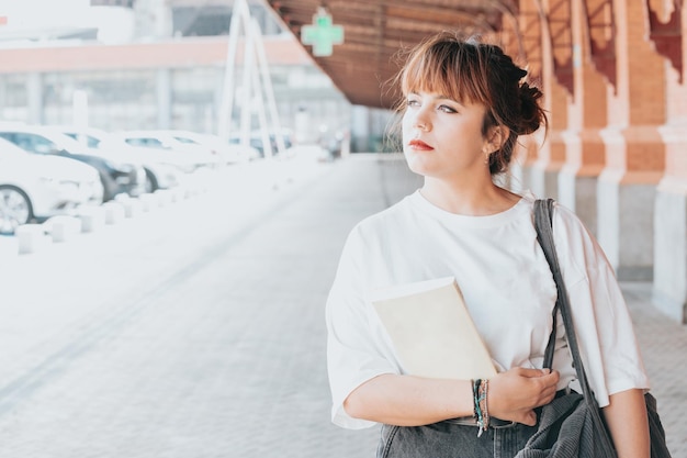 Młoda piękna czerwona głowa patrząc poważnie do kamery hipster kobiety. Trzymając książkę w pobliżu dworca kolejowego. Modna dziewczyna w letniej koszulce. zamyślona kobieta pozowanie na ulicy w pobliżu muru.