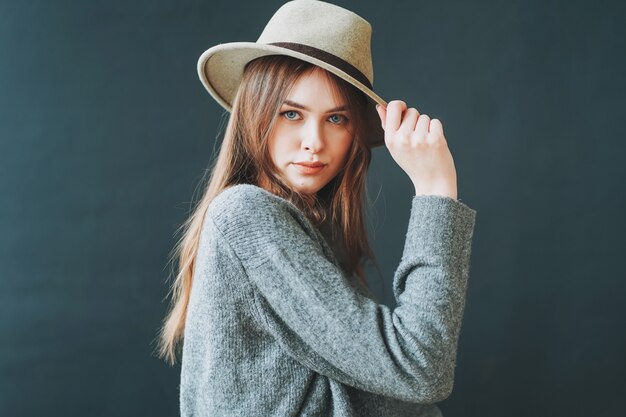 Młoda piękna długa brązowowłosa dziewczyna w filcowym kapeluszu i szarym swetrze z dzianiny, patrząc na kamery na ciemnym tle