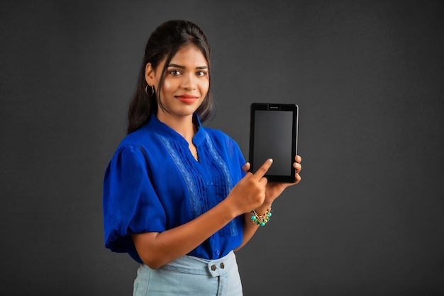 Młoda piękna dziewczyna pokazuje pusty ekran smartfona lub telefonu komórkowego lub tabletu na szarym tle