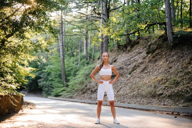 Młoda piękna dziewczyna pozuje przed treningiem biegowym, na drodze w gęstym lesie, podczas zachodu słońca. Zdrowy tryb życia i bieganie na świeżym powietrzu.