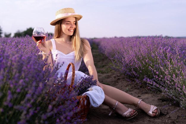 Młoda piękna dziewczyna siedząca wśród lawendowych krzaków latem o zachodzie słońca trzyma w rękach kieliszek z czerwonym winem. Degustacja wina
