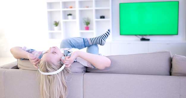 Młoda piękna dziewczyna słuchająca muzyki przez słuchawki, leżąca na kanapie w domu
