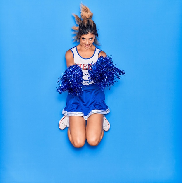 Młoda piękna dziewczyna ubrana w mundur cheerleaderki uśmiechnięta szczęśliwa Skakanie z uśmiechem na twarzy za pomocą pomponów na odizolowanym niebieskim tle
