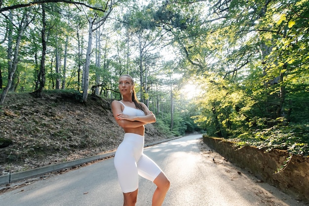 Młoda piękna dziewczyna w białych ubraniach pozuje przed treningiem biegowym na drodze w gęstym lesie podczas zachodu słońca Zdrowy styl życia i bieganie na świeżym powietrzu Młodzież