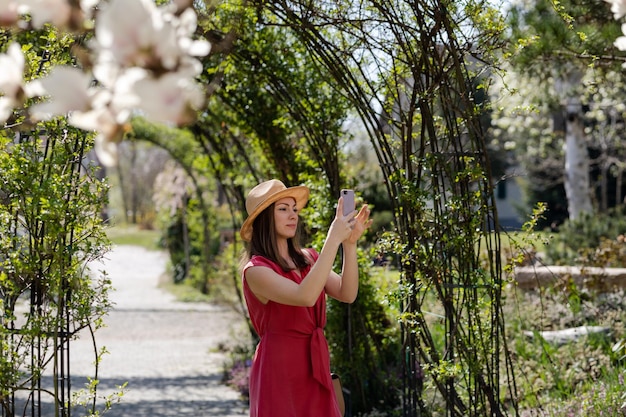 Młoda piękna dziewczyna w eleganckiej czerwonej sukience i słomkowym kapeluszu robi selfie na smartfonie na tle łuku róż i kwitnącej białej magnolii