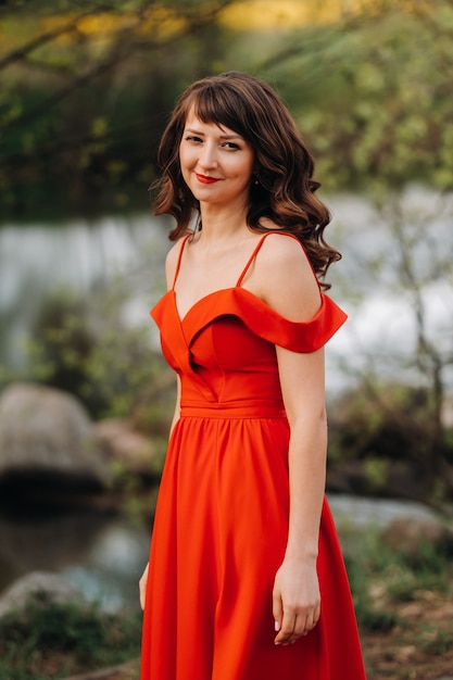 Młoda piękna dziewczyna z długimi brązowymi włosami, w długiej czerwonej sukience z pierścieniem wokół jeziora.