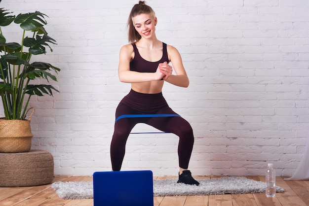 Młoda, piękna dziewczyna zajmuje się fitnessem w domu. Dziewczyna wykonuje różne ćwiczenia przy użyciu gumy fintes. Zawodnik jest szkolony przez Internet