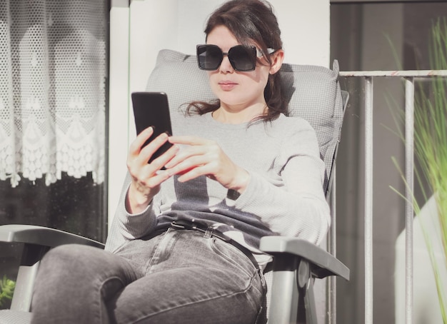 Młoda piękna kaukaska dziewczyna trzyma przed sobą smartfona, siedząc na krześle ogrodowym