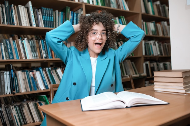 Młoda piękna kędzierzawa dziewczyna w szkłach i błękitnym kostiumu obsiadaniu z książkami w bibliotece.