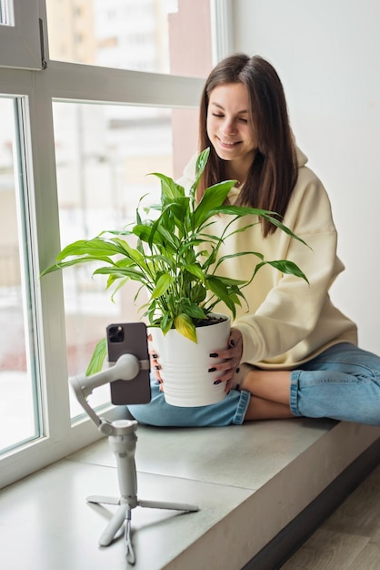 Młoda piękna kobieca blogerka blogująca o roślinach doniczkowych filmująca się z doniczkową spathiphyllum na smartfonie na statywie stabilizatora Miękkie selektywne ustawianie ostrości