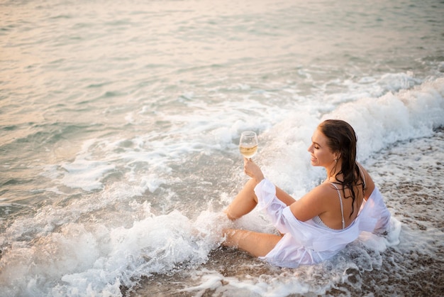 Młoda piękna kobieta nosi strój kąpielowy trzyma kieliszek wina siedząc w morskiej pianie fal nad naturą