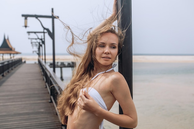 Młoda piękna kobieta o brązowych włosach w białym bikini elegancko pozuje na molo na tle plaży z piaskiem, oceanem i niebem. Tropikalna sesja zdjęciowa.