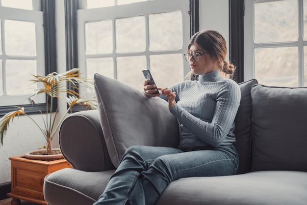 Młoda Piękna Kobieta Siedzi Na Kanapie W Domu, Rozmawiając I Surfując Po Sieci Kobieta Bawi Się Ze Smartfonem Online Portret Dziewczyny Uśmiecha Się Przy Użyciu Telefonu Komórkowego, Poprawiając Media Społecznościowe