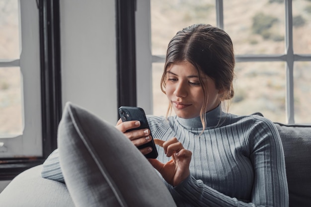 Młoda piękna kobieta siedzi na kanapie w domu, rozmawiając i surfując po sieci Kobieta bawi się ze smartfonem online Portret dziewczyny uśmiecha się przy użyciu telefonu komórkowego, poprawiając media społecznościowe