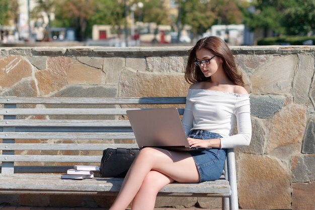 Młoda piękna kobieta siedzi na ławce w parku i pracuje na laptopie