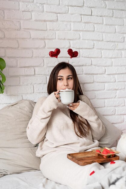Młoda piękna kobieta siedzi w łóżku świętując walentynki jedząc słodycze i pijąc kawę