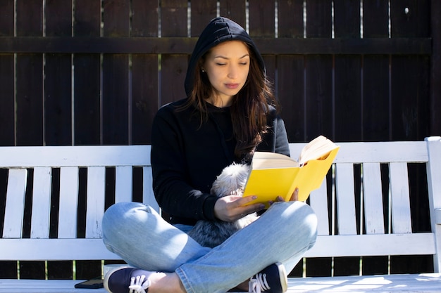 Młoda piękna kobieta siedzi ze zwierzakiem na drewnianej huśtawce na podwórku wiejskiego domu, czytając książkę