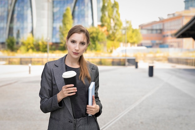 Młoda piękna kobieta stoi w garniturze na ścianie budynku biurowego. Pije kawę z filiżanki wielokrotnego napełniania, dbając o środowisko.