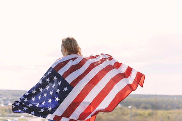 Młoda piękna kobieta trzymająca flagę USA Tył młodej kobiety z flagą narodową USA na ramionach na tle gór świętuje Dzień Niepodległości Stanów Zjednoczonych