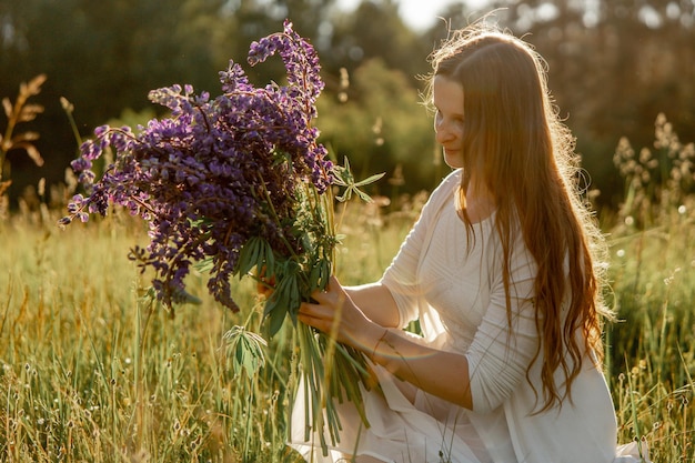 Młoda piękna kobieta ubrana w białą sukienkę trzymająca kwiaty na łące Dziewczyna ciesząca się naturą i wolnością Naturalne piękno Bezstresowe śnienie Zachód słońca