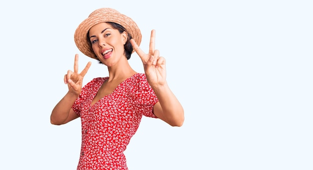 Młoda piękna kobieta ubrana w letni kapelusz, uśmiechnięta, patrząca w kamerę, pokazująca palce robiące znak zwycięstwa. numer dwa.