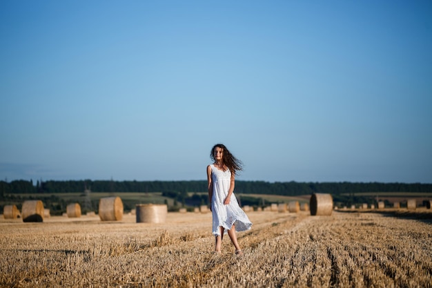 Młoda piękna kobieta w białej letniej sukience stoi na skoszonym polu pszenicy z ogromnymi snopami siana, ciesząc się przyrodą. Przyroda we wsi. Selektywne skupienie