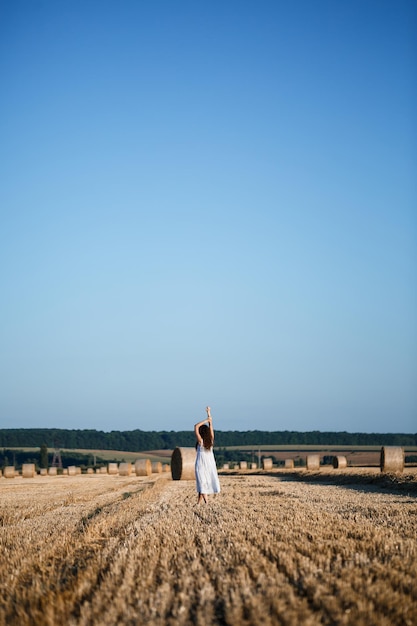 Młoda piękna kobieta w białej letniej sukience stoi na skoszonym polu pszenicy z ogromnymi snopami siana, ciesząc się przyrodą. Przyroda we wsi. Selektywne skupienie