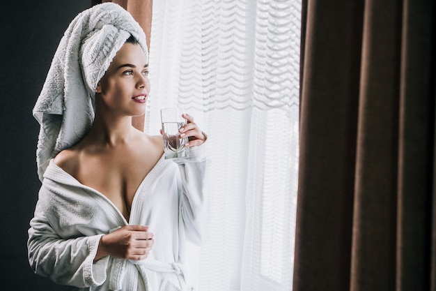 Młoda piękna kobieta w białym ręczniku i szlafroku, trzymając szklankę wody w pobliżu okna w domu