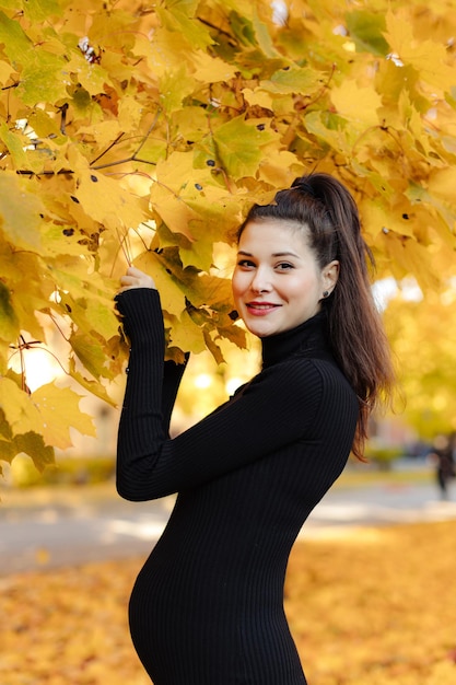 Młoda piękna kobieta w ciąży o ciemnych włosach w czarnej obcisłej sukience pozuje na jesień