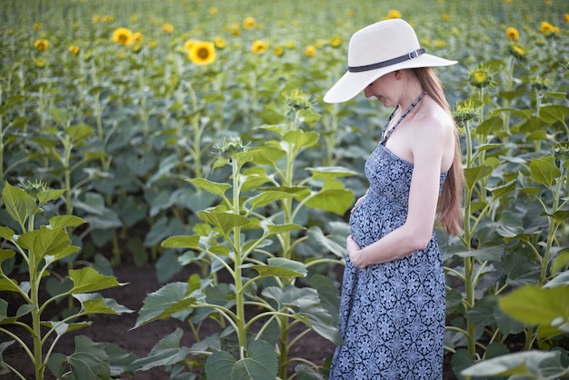 Młoda piękna kobieta w ciąży stoi w kapeluszu i sukni na polu kwitnących słoneczników