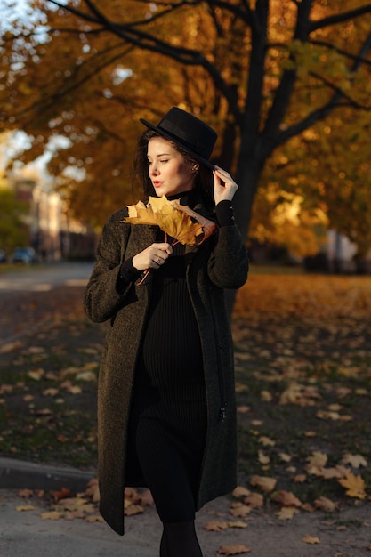 Młoda piękna kobieta w ciąży w kapeluszu z ciemnymi włosami w czarnej obcisłej sukience i płaszczu pozowanie
