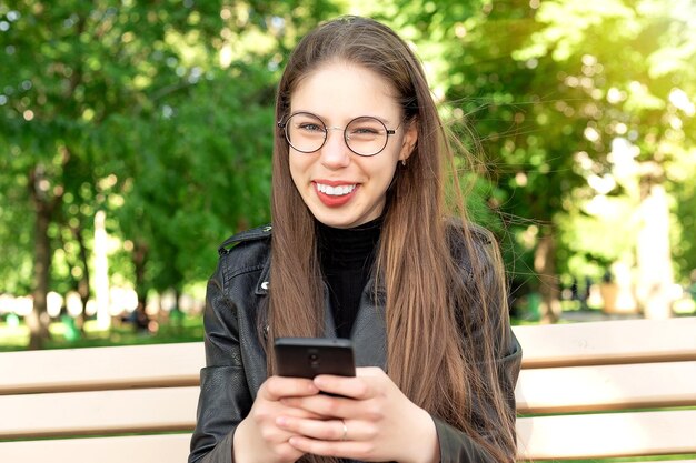 Młoda piękna kobieta w czarnej skórzanej kurtce pisze wiadomość lub gra w gry lub ogląda filmy lub wiadomości z mediów społecznościowych przez telefon, siedząc na ławce w pięknym zielonym letnim lub wiosennym parku