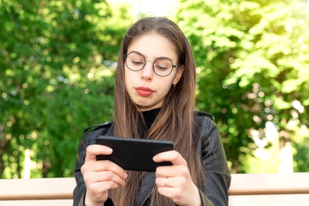 Młoda Piękna Kobieta W Czarnej Skórzanej Kurtce Pisze Wiadomość Lub Gra W Gry Lub Ogląda Filmy Lub Wiadomości Z Mediów Społecznościowych Przez Telefon, Siedząc Na ławce W Pięknym Zielonym Letnim Lub Wiosennym Parku