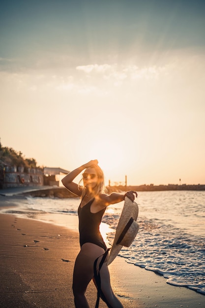 Młoda piękna kobieta w czarnym stroju kąpielowym i kapeluszu w okularach spaceruje po plaży o zachodzie słońca Koncepcja rekreacji na morzu Selektywna ostrość