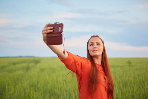 Zdjęcie młoda piękna kobieta w czerwonej sukience i rude włosy, bierze selfie na telefon w polu pszenicy zielony wieczorem