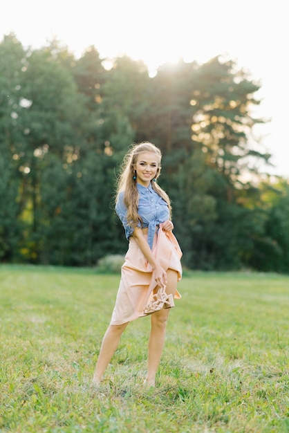Młoda piękna kobieta w letniej sukience przechodzi przez łąkę na zielonej trawie Młoda dziewczyna trzyma buty w rękach pokazując swoją piękną nogę Letnia rekreacja na świeżym powietrzu
