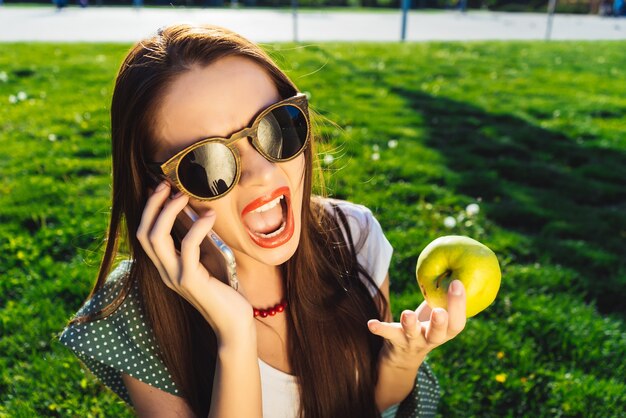 Młoda piękna kobieta w okularach przeciwsłonecznych siedzi na trawniku z jasnozieloną trawą, rozmawia przez telefon i krzyczy